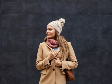 Winter fashion and woman style. A happy stylish woman in a coat, scarf, and beanie standing on the street and adjusting her coat while looking away.