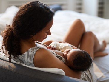 Peaceful loving young African mother sitting on bed and leaning on headboard while feeding baby with breast