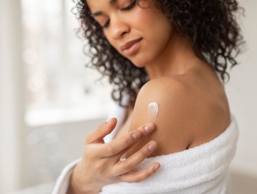 Skin care products. Young black lady applying moisturizing cream or lotion on body after shower, wearing bathrobe in bathroom, closeup, cropped