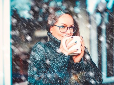Close-up portrait shot of smiling young woman holding a cup of tea in her hand and daydreaming while standing behind the window. It’s snowing outside.