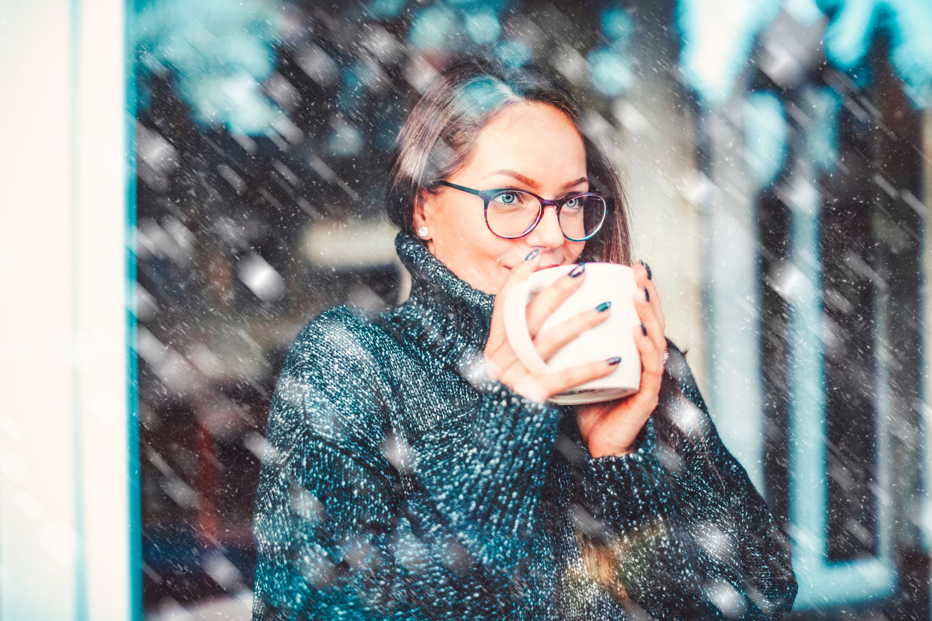 Close-up portrait shot of smiling young woman holding a cup of tea in her hand and daydreaming while standing behind the window. It’s snowing outside.
