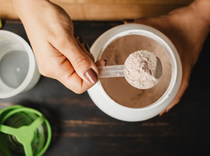 Female hand holds measuring spoon with portion of whey protein powder above plastic jar on wooden table with shaker, banana and apple fruit. Process of making protein drink, top view.