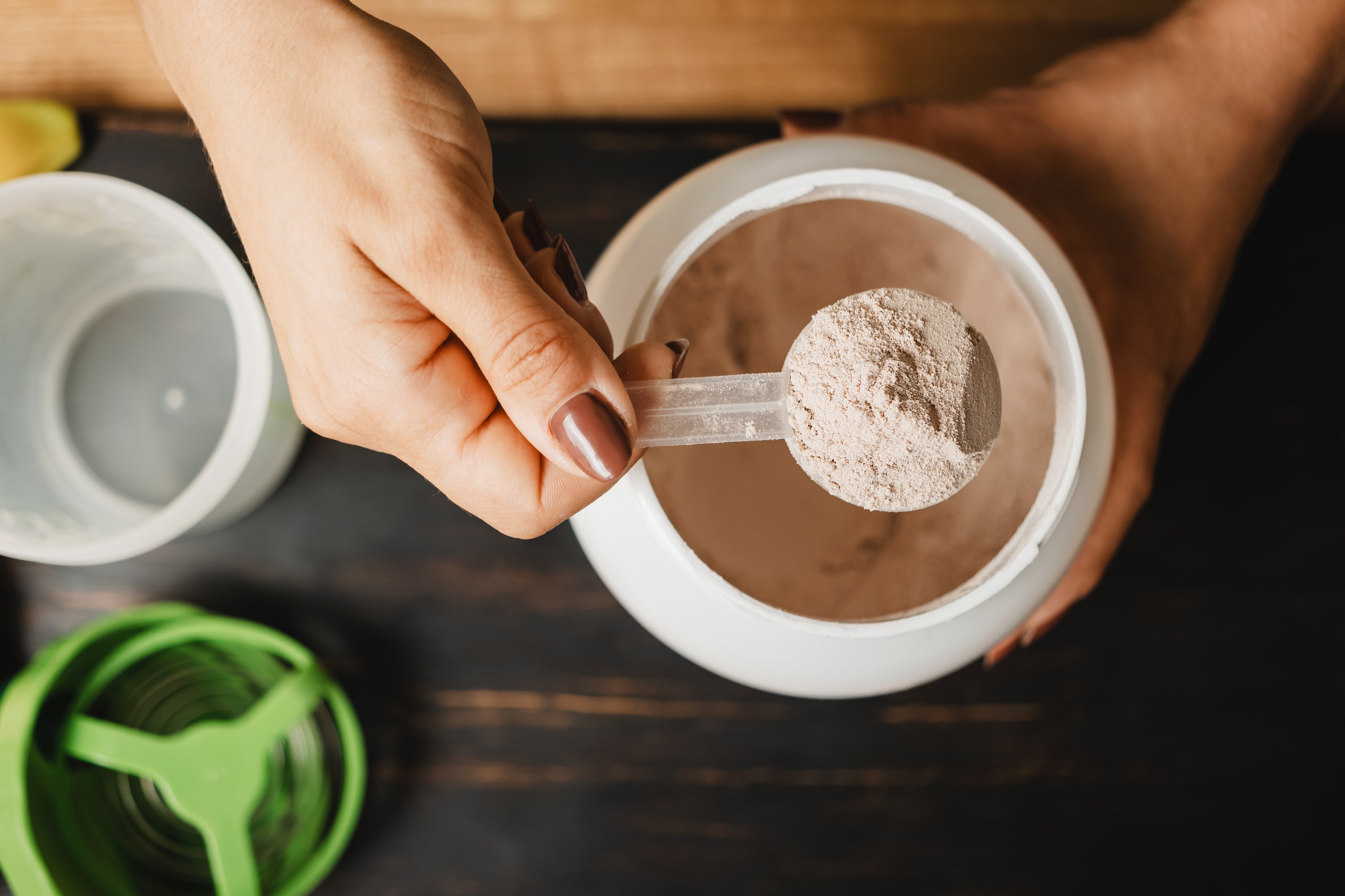 Female hand holds measuring spoon with portion of whey protein powder above plastic jar on wooden table with shaker, banana and apple fruit. Process of making protein drink, top view.