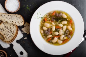 Healthy homemade vegetarian soup with white beans and mushrooms in white plate on gray background served with whole wheat bread viewed from above