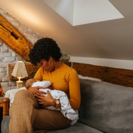 Black mother breastfeeding her baby while sitting on sofa at home