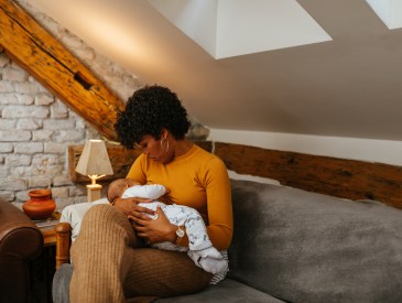 Black mother breastfeeding her baby while sitting on sofa at home