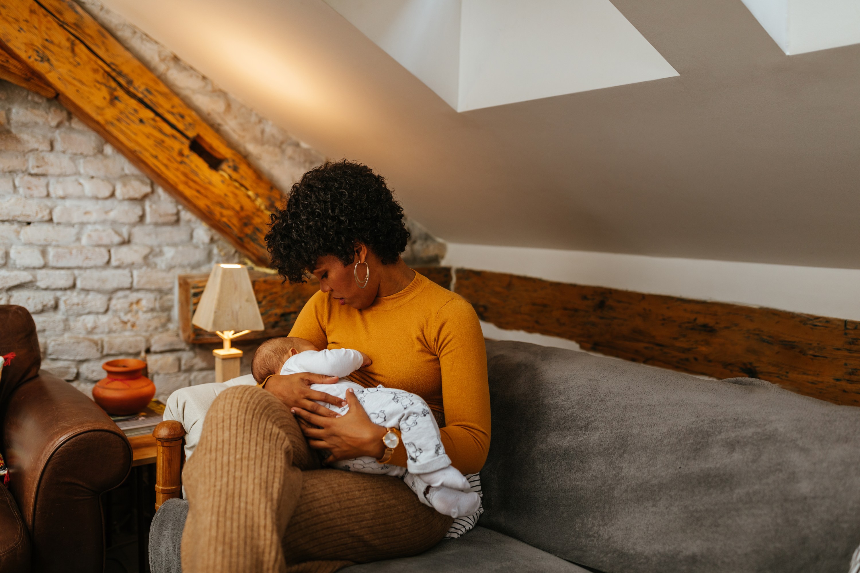 Black mother breastfeeding her baby while sitting on sofa at home